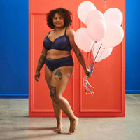 Femme ronde en lingerie chic tenant des ballons roses devant un mur coloré, mettant en avant la confiance et le style.