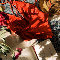 Maillot de bain rouge élégant posé sur un tissu, entouré de fleurs et d'un chapeau de paille.