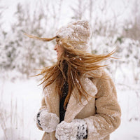 Femme souriante en manteau chaud, bonnet, et gants, se tenant dans la neige en hiver.